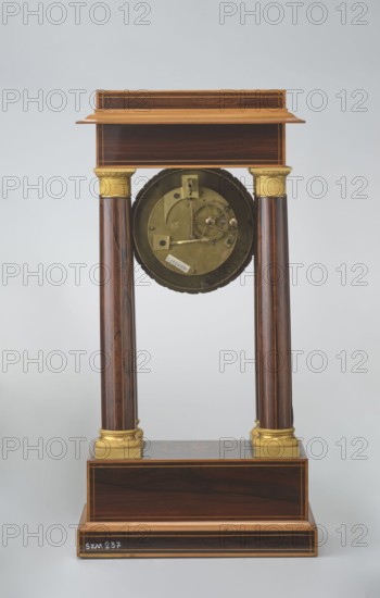 Mantel clock by Alexandre Pertrand fl. ca. 1840. Four-column portico form on a cuboid base and counter-base; boxwood inlays on the front of the cornice and base with a rhombus rosette and a tendril on both pilasters; gilded bronze bases and capitals. Suspended movement with a silver dial with Roman numerals and two keyholes hangs between the columns. Gilded bronze frame with shells surrounds the clock. Oak construction with rosewood facing and light wood insets. Circa 1840, Paris, France. Timepiece; height 47 cm, width 21 cm., A tall wooden structure with a rectangular base and a stepped top supported by a pair of polished vertical columns, each topped with a decorative gold-colored capital and resting on gold-colored bases, with a circular metallic mechanism suspended between the columns; the wood shows dark brown and lighter brown grain with thin lighter inlaid edges, the metallic parts are brass-colored and the capitals and bases are gold-colored, the background is pale gray.