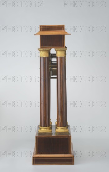 Mantel clock by Alexandre Pertrand fl. ca. 1840. Mantel clock in the form of a four-column portico on a cuboid base and counter-base; boxwood inlays on the front of the cornice and base with a rhombus rosette and a tendril on both stylons; gilded bronze bases and capitals. Suspended movement with a silver dial with Roman numerals and two keyholes hangs between the columns. Gilded bronze frame with shells surrounds the clock. Oak construction with rosewood facing and light wood insets. Ca. 1840, Paris, France. Timepiece; H. 47 cm, W. 21 cm., A vertical wooden structure with a polished dark brown finish and visible wood grain stands against a pale gray background; it has a rectangular stepped base and a small rectangular top shelf in a lighter brown, connected by a group of four tall cylindrical columns with dark brown wood and lighter brown streaks, each topped and bottomed with decorative gold-colored capitals and bases, and some exposed metallic mechanical components visible between t...