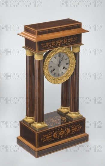 Mantel clock by Alexandre Pertrand fl. ca. 1840. Four-column portico form on a cuboid base and counter-base; boxwood inlays on the front of the cornice and base with a rhombus rosette and tendril on both stanchions; gilded bronze bases and capitals. Suspended movement with a silver dial with Roman numerals and two keyholes hanging between the columns. Gilded bronze frame with shells surrounds the clock. Oak construction with rosewood facing and light wood insets. Ca. 1840, Paris, France. Timepiece; height 47 cm, width 21 cm., A decorative wooden mantel clock with a rectangular base and top connected by four fluted columns, featuring a circular clock face framed by an ornate round gold-colored floral bezel; the wood shows dark brown and lighter brown tones with inlaid floral marquetry in a lighter wood color on the base and top, and gold-colored caps and square plinths at the column ends; the clock face is silver-gray with dark hands, set against a neutral pale gray background.