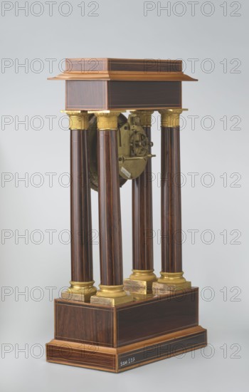 Mantel clock by Alexandre Pertrand fl. ca. 1840. Four-column portico form on a cuboid base and counter-base; boxwood inlays on the front of the cornice and base with a rhombus rosette and tendril on both styrons; gilded bronze bases and capitals. Suspended movement with a silver dial with Roman numerals and two keyholes hangs between the columns. Gilded bronze frame with shells surrounds the clock. Oak construction with rosewood facing and light wood insets. Ca. 1840, Paris, France. Timepiece; height 47 cm, width 21 cm., A freestanding wooden structure with a rectangular stepped base and a matching rectangular top supported by a group of tall, round columns; the columns have polished dark brown wood shafts with lighter brown wood grain and gilded capitals and bases, and there are gilded metal elements and a visible round mechanical component suspended between the columns; the wooden base and top show banded inlay in lighter brown tones, and the overall color palette includes dark brown...