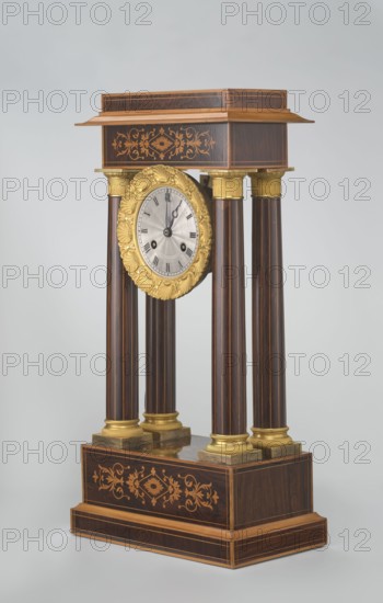 Mantel clock by Alexandre Pertrand fl. ca. 1840. Mantel clock in the form of a four-column portico on a cuboid base and counter-base; boxwood inlays on the front of the cornice and base with a rhombus rosette and a tendril on both stylons; gilded bronze bases and capitals. A suspended movement with a silver dial with Roman numerals and two keyholes hangs between the columns. A gilded bronze frame with shells surrounds the clock. Oak construction with rosewood facing and light wood insets. Ca. 1840, Paris, France. Timepiece; height 47 cm, width 21 cm., A tall decorative clock with a rectangular wooden top and base, connected by a group of four vertical cylindrical columns, each dark brown with lighter brown vertical streaks and gold-colored bases and capitals; a round clock face with a textured light silver center is set within an ornate gold-colored floral frame suspended between the upper support and the columns; the wooden surfaces feature inlaid floral and geometric marquetry in lig...