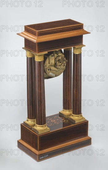 Mantel clock by Alexandre Pertrand fl. ca. 1840. Mantel clock in the form of a four-column portico on a cuboid base and counter-base; boxwood inlays on the front of the cornice and base with a rhombus rosette and a tendril on both stiles; gilded bronze bases and capitals. Suspended movement with a silver dial with Roman numerals and two keyholes hangs between the columns. Gilded bronze frame with shells surrounds the clock. Oak construction with rosewood facing and light wood insets. Ca. 1840, Paris, France. Timepiece. Height 47 cm, width 21 cm., A freestanding wooden structure with a rectangular top and base in dark brown wood with lighter brown trim, supported by a group of four tall fluted columns in dark brown with thin lighter streaks and gold-colored capitals and bases, an exposed circular brass mechanism suspended between the columns, and a dark brown plinth with a decorative floral inlay in lighter brown and gold tones, all set against a pale gray background.