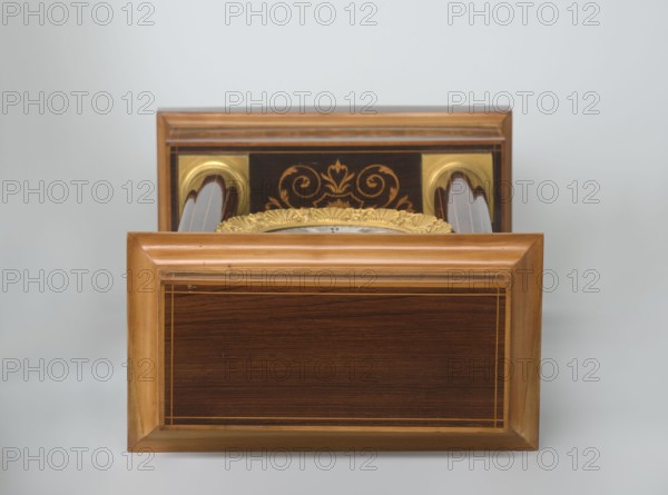 Mantel clock by Alexandre Pertrand fl. ca. 1840. Mantel clock in the form of a four-column portico on a cuboid base and counter-base; boxwood inlays on the front of the cornice and base with a rhombus rosette and a tendril on both pilasters; gilded bronze bases and capitals. A suspended movement with a silver dial with Roman numerals and two keyholes hangs between the columns. A gilded bronze frame with shells surrounds the clock. Oak construction with rosewood facing and light wood insets, ca. 1840, Paris, France. Timepiece; height 47 cm, width 21 cm., The image shows a wooden rectangular object with polished light brown and dark brown wood panels and a framed edge, set against a pale gray background; behind it is another similar wooden section with decorative gold-colored ornate detailing and curved elements partially visible, and a hint of a gold-colored decorative rim peeking above the front panel. The visible colors include light brown, dark brown, gold, pale gray, and a few darke...