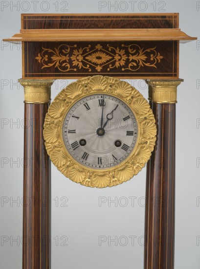 Mantel clock by Alexandre Pertrand fl. ca. 1840. Four-column portico form on a cuboid base and counter-base; boxwood inlays on the front of the cornice and base with a rhombus rosette and a tendril on both sturons; gilded bronze bases and capitals. Suspended movement with a silver dial with Roman numerals and two keyholes hanging between the columns. Gilded bronze frame with shells surrounds the clock. Oak construction with rosewood facing and light wood insets. Ca. 1840, Paris, France. Timepiece; height 47 cm, width 21 cm., A wooden clock structure with a rectangular top panel featuring ornate floral inlay in lighter brown tones above a gold-colored decorative round clock face rim, supported by a pair of dark brown cylindrical columns with lighter vertical streaks and gold-colored capitals; the round clock face has a silver-gray central disk with darker hands and a textured pattern, and the overall visible colors include dark brown, light brown, gold, silver-gray, and black.