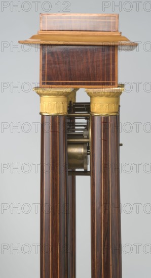 Mantel clock by Alexandre Pertrand fl. ca. 1840. Mantel clock in the form of a four-column portico on a cuboid base and counter-base; boxwood inlays on the front of the cornice and base with a rhombus rosette and a tendril on both styrons; gilded bronze bases and capitals. A suspended movement with a silver dial with Roman numerals and two keyholes hangs between the columns. A gilded bronze frame with shells surrounds the clock. Oak construction with rosewood facing and light wood insets. Circa 1840, Paris, France. Timepiece. Height 47 cm, width 21 cm., A vertical section of an object showing a wooden upper block with a smooth, polished dark brown surface and lighter brown trim, topped by a slightly overhanging cap in a medium golden-brown color. Below the block are a pair of decorative cylindrical capitals in a metallic gold color with embossed detailing, supporting a set of long, slender columns with a glossy dark brown wood grain and thin lighter brown vertical stripes. Between the ...