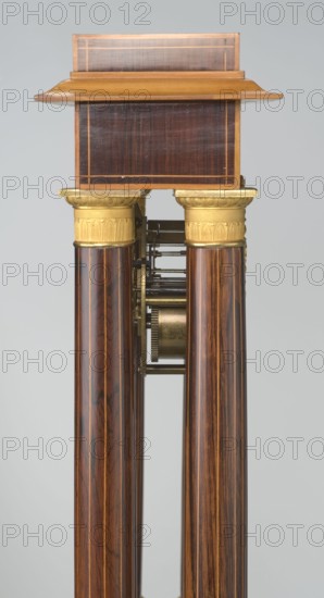 Mantel clock by Alexandre Pertrand fl. ca. 1840. Four-column portico form on a cuboid base and counter-base; boxwood inlays on the front of the cornice and base with a rhombus rosette and tendril on both sides; gilded bronze bases and capitals. Suspended movement with a silver dial bearing Roman numerals and two keyholes hangs between the columns. Gilded bronze frame with shells surrounds the clock. Oak construction with rosewood facing and light wood insets. Ca. 1840, Paris, France. Timepiece; height 47 cm, width 21 cm., The image shows a tall, narrow wooden structure with a rectangular top piece in dark brown wood framed by lighter brown edges and a slightly overhanging shelf in a warm golden-brown tone, supported by a pair of cylindrical columns in richly grained dark brown wood with lighter streaks; the tops of the columns have ornate gold-colored capitals with decorative motifs and a gold-colored mechanical assembly is visible between the columns; the background is a plain light g...