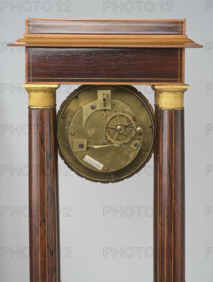 Mantel clock by Alexandre Pertrand fl. ca. 1840. Mantel clock in the form of a four-column portico on a cuboid base and counter-base; boxwood inlays on the front of the cornice and base, with a rhombus rosette and a tendril on both stiles; gilded bronze bases and capitals. A suspended movement with a silver dial with Roman numerals and two keyholes hangs between the columns. A gilded bronze frame with shells surrounds the clock. Oak construction with rosewood facing and light wood insets. Ca. 1840, Paris, France. Height 47 cm; width 21 cm., An upright wooden structure with a rectangular top section in dark brown wood and lighter brown trim, supported by a pair of polished cylindrical wooden columns showing rich brown and reddish wood grain with darker streaks; each column has a decorative gold-colored capital near the top. Between the columns hangs a round metallic mechanism in a muted brass color with visible gears and small fastenings, and a small pale rectangular label attached to i...