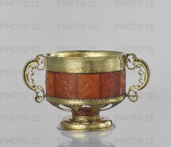 Platter with amber plates, cylindrical platter on a slightly arched circular foot with oval cutouts at the bottom; central amber plates decorated with flowers; upper edge enclosed in a silver hoop engraved with leafy tendril motifs; scrollwork handles with pearling on the sides; 17th century, possibly Królewiec; vessel., A decorative bowl with ornate scrolled handles sits against a plain light gray background; the bowl features a band of warm reddish-brown panels around its midsection with carved floral motifs, framed by a gleaming yellow-gold metal rim and base with engraved patterns, and the handles are filigreed and beaded in the same yellow-gold color, with openwork details showing glimpses of the background through the lower base.