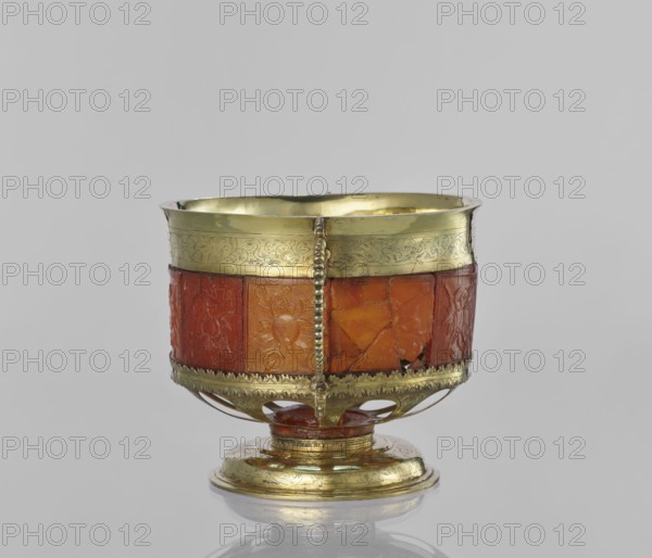 Platter with amber plates, cylindrical platter on a slightly arched circular foot with oval cutouts at the bottom; central amber plates decorated with flowers; upper edge enclosed in a silver hoop engraved with leafy tendril motifs; sides with scrollwork handles with pearling; garlands and scrollwork ornament; 17th century, Królewiec, vessel;, A decorative cup-like object sits centered against a plain light gray background, showing a rounded bowl mounted on a short pedestal base. The upper rim and base appear metallic with a reflective yellow-gold color and engraved ornamental patterns, while the middle band of the bowl is composed of carved panels in warm orange and reddish-orange tones with floral and geometric motifs. A vertical beaded strip of the same yellow-gold metal runs down the front, connecting the rim to the lower decorative edge, and openwork elements in the metal form arches above the pedestal. The interior of the bowl shows the same yellow-gold reflective surface, and th...