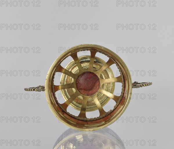Platter with amber plates, cylindrical platter on a slightly arched circular foot with oval cutouts at the bottom; central amber plates decorated with flowers; upper edge enclosed in a silver hoop engraved with leafy tendril motifs; scrollwork handles with pearling on the sides; 17th century, possibly Królewiec; vessel., A circular metallic object viewed from the front with a ringed outer rim and radial spokes converging on a central disk, showing metallic gold tones and areas of reddish-brown patina, a few ornate handles extending outward on either side with darker metallic details, and a pale gray background with a reflective surface beneath that creates a faint mirrored image.
