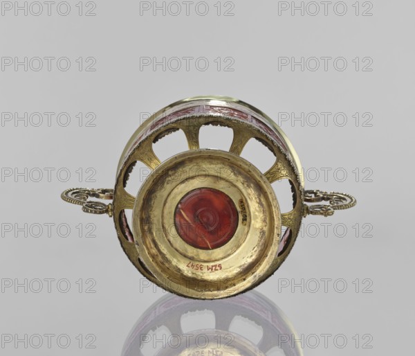 Platter with amber plates, cylindrical platter on a slightly arched circular foot with oval cutouts at the bottom; central amber plates decorated with flowers; upper edge enclosed in a silver hoop engraved with leafy tendril motifs; scrollwork handles with pearling; 17th century, Królewiec possible place of origin; vessel., A circular metal object with a patinated gold surface and ornate openwork sides is shown laying on its rim, featuring a central round inset of translucent deep red glass; the metal has engraved decorative patterns and a pair of twisted rope-style handles extending horizontally from each side, and the scene includes reflections of the object on a glossy gray surface beneath; visible colors include gold, red, gray, and a hint of purple in the reflected area.