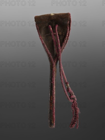 Baroque spoon case with the Korczak coat of arms, coats of arms, Korczak coat of arms, baroque style. Leather cover embroidered with metal thread showing the Korczak coat of arms and SL monogram, tied with crimson cords. Very few cases of this type have been found; this one, along with the spoon for which it was made, is particularly rare. 1st half of the 18th century, Poland, possibly Lviv. Cutlery., A long narrow object with a tapered handle and a wider top, covered in worn brown material with visible texture, has a group of braided cords attached that are a faded reddish-purple color with some lighter threads, ending in frayed tassels; the edges of the top have reddish-purple stitching, and there are small areas of lighter brown and a touch of yellowish discoloration on the surface, all set against a neutral gray background.