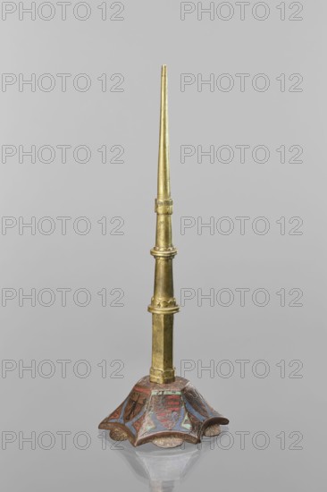 Candlestick, enamel; on a hexagonal base tapering toward the top with a shaft; on each side a coat of arms on a blue enamel background eagle and blue lion on horizontal red stripes; between them below the shields on the edges rhombuses with engraved dragons. 19th-century shaft, 14th century Limoges, Barcelona, lighting; height 32 cm, diameter 11 cm., A slender vertical object with a pointed top in a metallic gold color rises from a decorative base; the shaft shows a few segmented collars and cylindrical sections in varying gold tones with subtle darker patina. The base is flared and scalloped with a curved, petal-like shape, painted in reds, blues, and touches of teal with areas of brownish wear and faded pigments, and it rests on a reflective gray surface that mirrors part of the base. The background is a uniform light gray.