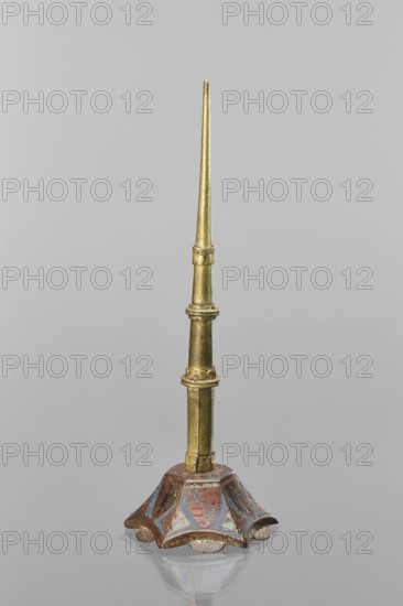 Candlestick, enamel on a hexagonal base tapering toward the top with a shaft; on each side a coat of arms on a blue enamel background eagle and blue lion on horizontal red stripes; between them, below the shields on the edges, rhombuses with engraved dragons. 19th-century shaft; 14th-century Limoges possibly; Barcelona possibly; lighting; height 32 cm, diameter 11 cm., A tall, slender metallic object with a tapered golden shaft rising from a decorative base; the shaft shows a few segmented rings and slight ornamentation and is a metallic gold color. The base is flared and scalloped with a worn painted surface showing muted reds, blues, and hints of green alongside the gold, and it sits on a reflective gray surface that mirrors the base faintly; the background is a uniform pale gray.