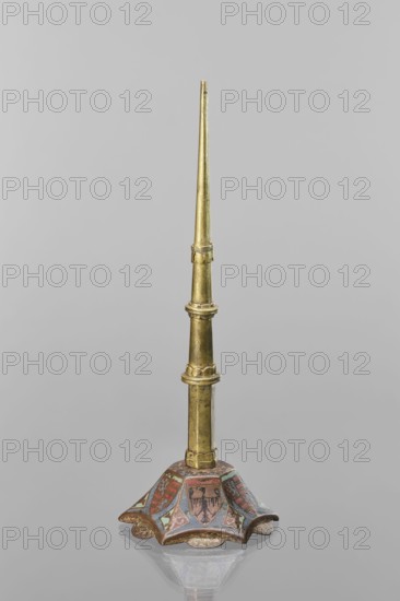 Candlestick, enamel, on a hexagonal base tapering towards the top with a shaft; on each side a coat of arms on a blue enamel background eagle and blue lion on horizontal red stripes, between them below the shields on the edges rhombuses with engraved dragons. 19th-century shaft, 14th century Limoges, Barcelona, lighting, height 32 cm, diameter 11 cm, A tall, slender, vertically oriented metallic object with a tapered shaft in a golden color rising from a decorative flared base; the shaft shows a few segmented rings and slight surface irregularities in the same golden tone. The base flares outward in a scalloped shape and features painted decorative panels in muted colors including red, blue, green, and brown, with repeating heraldic-style motifs and worn areas where the paint has aged. The object sits on a smooth reflective surface that creates a faint mirrored reflection beneath it, and the background is a uniform light gray.