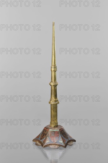 Candlestick, enamel on a hexagonal base tapering toward the top with a shaft; on each side a coat of arms on a blue enamel background eagle and blue lion on horizontal red stripes, between them below the shields on the edges rhombuses with engraved dragons. 14th century Limoges, and 19th-century shaft; lighting. Height 32 cm, diameter 11 cm., A tall, slender metallic object with a tapered upper section in a golden color rises from a decorative base; the base is flared and scalloped with a painted surface showing areas of red, blue, and brown, and the metal shows a slightly aged, patinated appearance; the background is a uniform light gray.