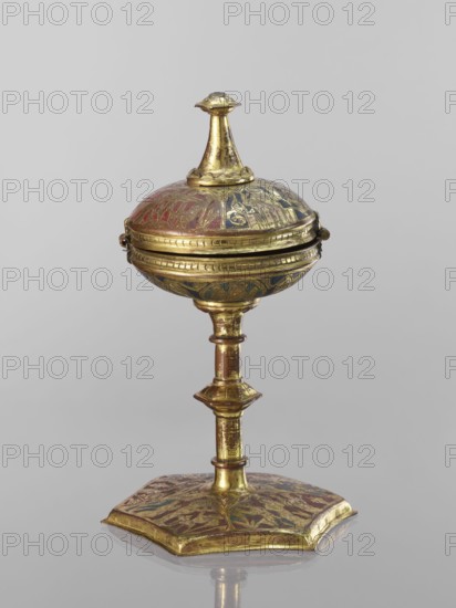 Host ciborium, Copper ciborium on a hexagonal foot, with a smooth shaft with two lacings in the form of a half-cylinder and a hexagonal nodus. The body is a flattened sphere, the upper half serving as a cover; the cover has a conical pinnacle originally topped with a cross. On the foot in the arcades are depictions of the Flagellation, Carrying the Cross, Crucifixion, and Christ in Majesty. On the bowl are busts of angels in circular medallions; on the cover in the trefoil arcades are four depictions of jhakas on the foot. Champlevé enamel in brick red and dark blue, late 14th century, Spain, ritual object; height 28 cm, diameter 16 cm., A gilded metal object with a round, domed top and a finial on its peak, mounted on a slender, segmented stem that rises from a flat, hexagonal base; the surface shows areas of worn gold with decorative painted or inlaid motifs in red, blue, and green, and the piece casts a soft reflection on a neutral gray background.