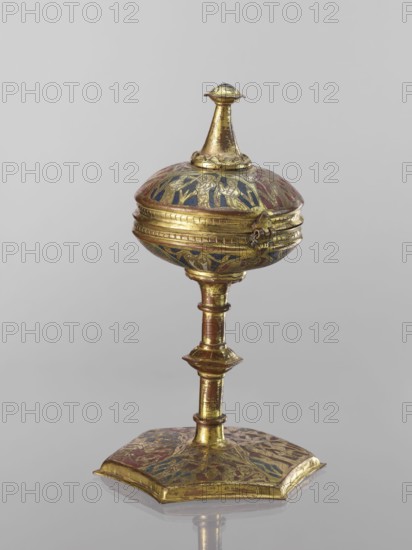 Host ciborium. Copper ciborium on a hexagonal foot, with a smooth shaft with two lacings in the form of a half-cylinder and a hexagonal nodus. The body is a flattened sphere, the upper half serving as a cover; on it is a conical pinnacle originally topped with a cross. In the arcades on the foot are depictions of the Flagellation, Carrying the Cross, Crucifixion, and Christ in Majesty. On the bowl are busts of angels in circular medallions; on the cover in trefoil arcades are four additional depictions. Champlevé enamel in brick red and dark blue. Late 14th century, Spain, ritual object. Height 28 cm, diameter 16 cm., A gilded metal object with an ornate rounded top and a flared finial sits on a slender, segmented stem that rises from a flat, hexagonal base; the surface shows areas of polished gold with decorative inlaid sections in blue and red, and engraved linear and floral motifs across the lid, stem, and base, with a small hooked clasp on the side of the rounded top.