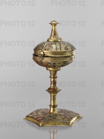 Host ciborium, Copper ciborium on a hexagonal foot, with a smooth shaft with two lacings in the form of a half-cylinder and a hexagonal nodus. The body is a flattened sphere, the upper half serving as a cover; the cover has a conical pinnacle originally topped with a cross. In the arcades on the foot are depictions of the Flagellation, Carrying the Cross, Crucifixion, and Christ in Majesty. The bowl bears busts of angels in circular medallions; the cover in trefoil arcades has four depictions jhakas. Champlevé enamel in brick red and dark blue. Late 14th century, Spain, ritual object. Height 28 cm, diameter 16 cm., A small ornate object with a rounded, lidded top mounted on a slender stem and a broad, shaped base, all shown against a plain light gray background. The object appears metallic with a dominant gold color and visible areas of red and blue decorative enamel or paint. The lid has a finial at the top and a hinge on the side, and the rounded body shows engraved or raised decorat...
