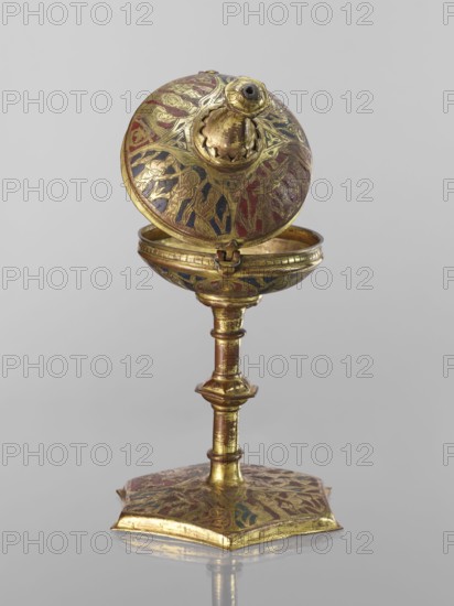 Host ciborium, Copper ciborium on a hexagonal foot, with a smooth shaft with two lacings in the form of a half-cylinder and a hexagonal nodus. The body is a flattened sphere, the upper half serving as a cover; on it is a conical pinnacle originally topped with a cross. In the arcades on the foot are depictions of the Flagellation, Carrying the Cross, Crucifixion, and Christ in Majesty; on the bowl are busts of angels in circular medallions; on the cover in the trefoil arcades are four depictions jhakas. Champlevé enamel in brick red and dark blue. Late 14th century, Spain, ritual object. Height 28 cm, diameter 16 cm., A decorative metal object stands against a plain light gray background, consisting of a rounded top that is hinged open to reveal an inner cavity, a slender columnar stem with ornamental rings and a square scalloped base, all showing areas of worn gilding. The surface of the top, inner bowl, stem, and base display intricate engraved and embossed patterns filled with patch...