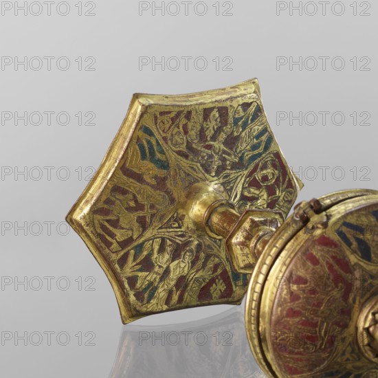 Host ciborium, Copper ciborium on a hexagonal foot, with a smooth shaft with two lacings in the form of a half-cylinder and a hexagonal nodus. The body is a flattened sphere, the upper half serving as a cover; the cover has a conical pinnacle originally topped with a cross. On the foot in arcades are depictions of the Flagellation, Carrying the Cross, Crucifixion, and Christ in Majesty. On the bowl are busts of angels in circular medallions; on the cover in trefoil arcades are four depictions. Champlevé enamel brick red and dark blue. Late 14th century, Spain, ritual object. Height 28 cm, diameter 16 cm., A close-up view of a metallic object with a hexagonal decorative element and an adjoining circular component, both showing engraved figural and vegetal patterns highlighted in gold tones with areas of red and blue enamel, and a central shaft connecting the pieces in a worn gold finish; the background is a smooth light gray and the object casts a subtle reflection on the surface beneat...