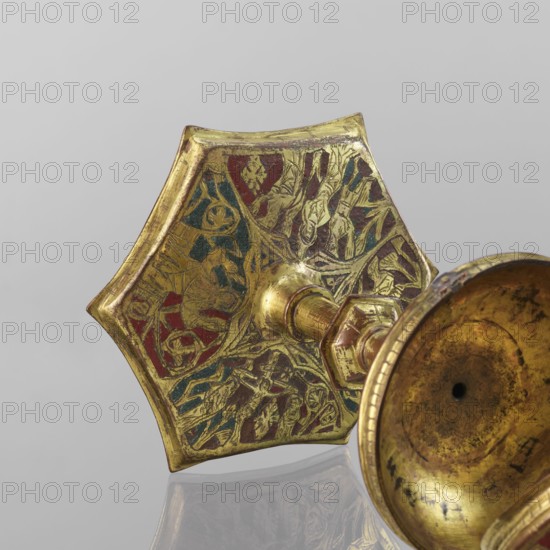 Host ciborium, Copper ciborium, on a hexagonal foot, with a smooth shaft with two lacings in the form of a half-cylinder, with a hexagonal nodus. The body is a flattened sphere, the upper half serving as a cover. On the cover is a conical pinnacle, originally topped with a cross. In the arcades on the foot are depictions of the Flagellation, Carrying the Cross, Crucifixion, and Christ in Majesty. On the bowl are busts of angels in circular medallions; on the cover in trefoil arcades are four depictions possibly of jhakas. Champlevé enamel, brick red and dark blue. Late 14th century, Spain . Ritual object. Height 28 cm, diameter 16 cm., The image shows a close-up of a decorative metallic object with a hexagonal base and a stem attached to a shallow bowl-like section; the metal has a worn gold finish with areas of red and blue enamel inlaid into engraved patterns, and the surface displays scratches and tarnish.