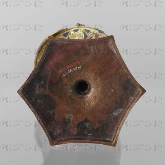 Host ciborium, Copper ciborium on a hexagonal foot, with a smooth shaft featuring two lacings in the form of a half-cylinder and a hexagonal nodus. The body is a flattened sphere, the upper half serving as a cover; the cover has a conical pinnacle originally topped with a cross. On the foot in the arcades are depictions of the Flagellation, Carrying of the Cross, Crucifixion, and Christ in Majesty. On the bowl are busts of angels in circular medallions; on the cover in the trefoil arcades are four depictions of jhakas on the foot. Champlevé enamel, brick red and dark blue, late 14th century, Spain, ritual object, height 28 cm, diameter 16 cm., A close-up view of the bottom of a hexagonal metal object with a central circular hole, showing a worn, textured surface in shades of brown, reddish-brown, dark brown, and hints of black, with areas of lighter tan and small patches of greenish corrosion; the object has a slightly raised rim and beveled edges, and a glimpse of an inner rim decorat...