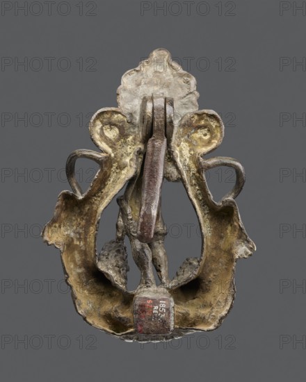 Bronze door knocker with a figure of Bacchus flanked by two lions with raised torsos. Renaissance style, attributed to the circle of Niccolò Roccatagliata Roccatagliara, late 16th–early 17th century, Venetian. Door fitting, height 22 cm, width 16 cm. Inscription/stamp: Dzialynska, Izabella 1830–1899. Variation exists showing an old man center and different top/bottom details; comparable examples include a knocker with Neptune from early 17th-century Venice., An aged metallic object with an ornate, symmetrical shape sits against a plain gray background; its surface shows a mix of dull brown, greenish, and yellowish tones with areas of darker brown and spots of lighter, almost whitish corrosion; the form includes a central vertical bar and a rounded lower section with a hollowed interior where a small sculptural element with textured, darker brown tones is partially visible; the outer edges have flowing, organic curves and scalloped details, and the piece overall displays a worn, uneven ...