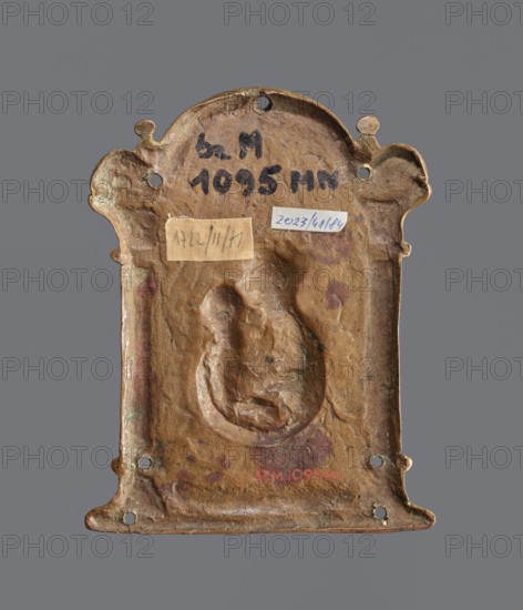 Pax pacific, Dzialynska, Izabella née Czartoryska 1830–1899, Goluchów ordination, enamel, liturgies. Plaque in the form of a frame with two columns topped by a semicircle, with a depiction of the Virgin Mary and Child; in the arch a depiction of the Crucified. White, blue, red, and green enamel. 16th century, Spain. Plaque; height 14.5 cm, width 11 cm., A small rectangular metal plaque with a rounded top and ornamental curved corners, shown against a gray background, with a raised central relief depicting a seated figure holding a smaller figure; the metal surface is weathered with a patina showing shades of brown, green, and reddish tones, and there are a few small round holes near the edges and a couple of small rectangular patches of lighter-colored material affixed to the surface.