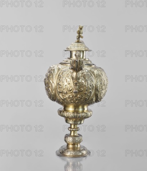Water tincture, Schweiger, Matthias fl. ca. 1623–1635 goldsmith, herms decorative elements, lions, flowers ornament, fruit ornament, acanthus ornament. Jug with a spherical belly decorated with repoussé bouquets of flowers and fruit in acanthus reserves; on the side a spout in the shape of a female half-figure holding a conch; on the axis a plastic handle formed of channel sections connected by a winged herm of a woman. The cover is topped with a plastic lion standing on its hind legs. 1630–1635, Augsburg, Germany. Vessel; height 28 cm, width 14 cm, depth 30 cm., A single ornate metal vessel with a bulbous body set on a pedestal base, featuring intricate raised floral and scrollwork patterns all over its surface and a small sculpted figure or decorative finial on the lid; the object shows a reflective metallic finish in shades of gold and brass, with highlights and subtle darker areas from shadows, sitting against a plain light gray background.