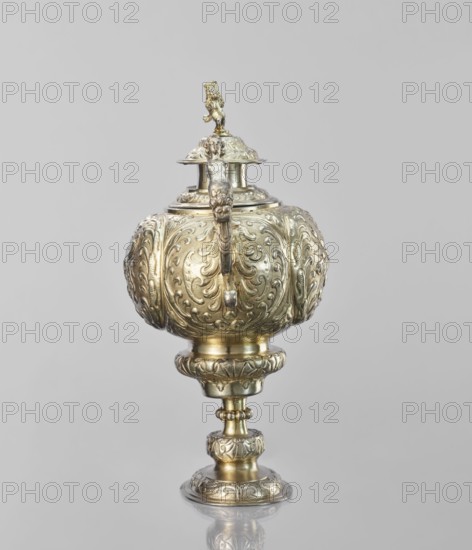 Water tincture, Schweiger, Matthias fl. ca. 1623–1635 goldsmith; herms decorative elements; lions; flowers ornament; fruit ornament; acanthus ornament. Jug with a spherical belly decorated with repoussé bouquets of flowers and fruit in acanthus reserves; on the side a spout in the shape of a female half-figure holding a conch; on the axis a plastic handle formed of channel sections connected by a winged herm of a woman. The cover is topped with a plastic lion standing on its hind legs. 1630–1635, Augsburg, Germany. Vessel; height 28 cm, width 14 cm, depth 30 cm., An ornate metal vessel with a rounded, bulbous body sits on a decorative pedestal; the vessel is covered in raised swirling and floral patterns and has a curved handle attached to its side, a domed lid topped with a small sculpted figure, and a reflective surface showing shades of gold and silver against a plain pale gray background.
