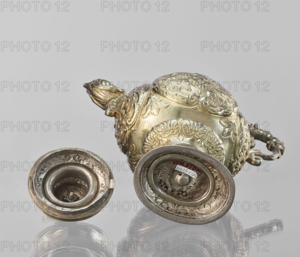 Water tincture, Schweiger, Matthias fl. ca. 1623–1635 goldsmith, herms decorative elements, lions, flowers ornament, fruit ornament, acanthus ornament. Jug with a spherical belly decorated with repoussé bouquets of flowers and fruit in acanthus reserves; on the side a spout in the shape of a female half-figure holding a conch; on the axis a plastic handle formed of channel sections connected by a winged herm of a woman. The cover is topped with a plastic lion standing on its hind legs. 1630–1635, Augsburg, Germany. Vessel; height 28 cm, width 14 cm, depth 30 cm., A metallic ornamental vessel and two matching lids lie on a reflective surface; the vessel shows ornate embossed floral and scrollwork patterns and a curved handle with decorative detailing, and the lids display similar embossed designs with concave inner areas. The visible colors are gold, silver, and shades of gray, with the background and reflections appearing light gray.
