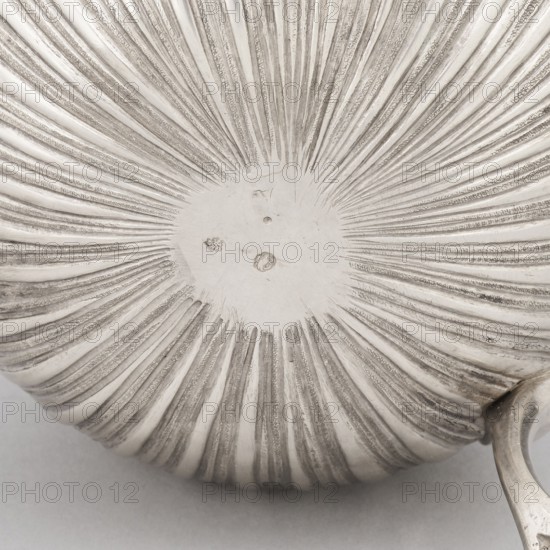 Chocolate jug, maker: Edme-Pierre Balzac fl. 1739–1781, goldsmith. Pear-shaped chocolate jug supported on three legs, with a small spout and a hinged lid; circular opening with a sliding circular cover. Lower part of the belly decorated with vertical curls; upper part with repoussé garlands and an oval reserve. Materials/technique: metalwork vieux Paris, France, Paris, dated circa 1764–1765. Vessel; height 26 cm, width 24 cm, depth 13.5 cm. Purchased in 1883 for 500 francs., A close-up view of a round, ridged metallic surface with a radial pattern emanating from a smoother central area, showing a group of concentric, textured grooves and raised lines; visible colors include shades of silver, gray, and off-white, with slight darker gray accents in the grooves and a small curved metallic handle partially visible at the lower right edge.