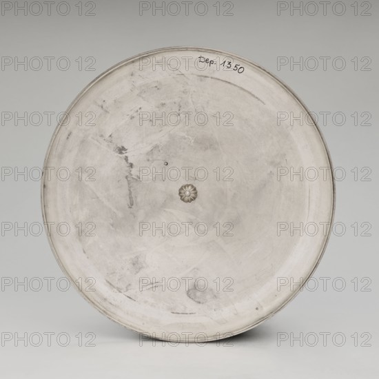 Empire vase by Jean-Baptiste-Claude Odiot goldsmith, co-created by Odiot, Maison goldsmith's firm. Empire style tableware vase, 1819–1838. Paris, France. Vessel; height 38.6 cm, width 39.0 cm., A large circular object with a smooth, slightly worn surface in shades of gray and off-white fills the image, showing subtle darker gray smudges and scuff marks across its face and a small raised, flower-shaped knob near the center with a slightly darker gray tone, all set against a pale gray background.
