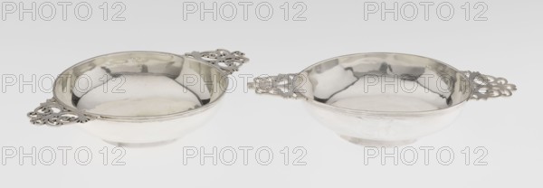 Pair of porridge bowls porringers by Wakely & Wheeler Ltd, London goldsmiths, fl. 1791–1958. Porringer, openwork, floral ornaments; plain bowls on circular plinths with a rim decorated by a profiled band and flat, triangular openwork handles on the sides, decorated with floral motifs. 1904–1905. England, London, Great Britain. Dish. Height 3.5 cm; width 20 cm; depth 22.8 cm; weight 387 g 1: 198.67 g; 2: 188.36 g., A pair of shallow round metal bowls with ornate pierced handles sit side by side on a light background; the bowls have reflective surfaces showing soft highlights and shadows and appear in shades of silver and gray with subtle warm reflections, and the decorative handles feature intricate filigree shapes in the same metallic color.