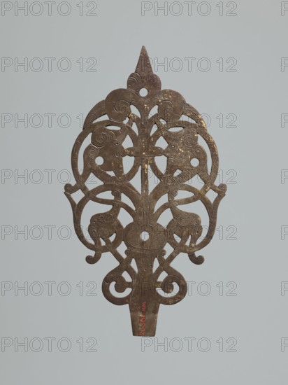 Hinge end for a chest, fittings, hinge end from a pair, in the form of an openwork rounded plaque with a tendril motif, two male masks among the twists. Hinge cut off; perhaps originally a disc hinge with a sleeve at the end of the plate or a strap hinge in the form of a clock hand. 1st half of the 17th century, Poland, Central Europe. Door fittings. Height 21.5 cm, width 12.5 cm, depth 0.2 cm., A vertically oriented decorative metal object with an openwork design featuring symmetrical, curving lines and scroll motifs, including stylized animal head shapes and rounded cutouts, rendered in brown and bronze tones against a pale gray background.