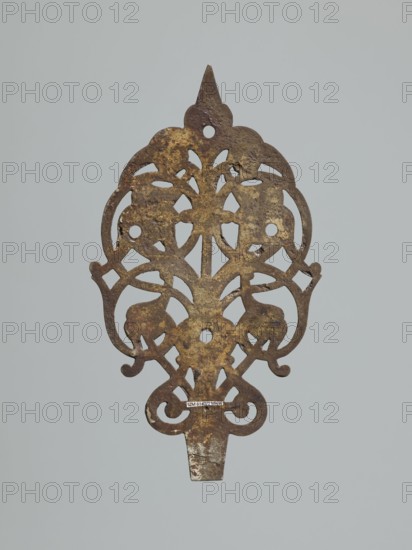 hinge end for a chest, fittings, hinge end from a pair, in the form of an openwork rounded plaque with a tendril motif, two male masks among the twists. Hinge cut off; perhaps originally it was a disc hinge with a sleeve at the end of the plate or a strap hinge, in the form of a clock hand, 1st half of the 17th century, Poland, Central Europe, door fittings, height 21.5 cm, width 12.5 cm, depth 0.2 cm, An ornate, flat metal object with an openwork, symmetrical design is shown against a pale gray background; the metal surface is mottled with brown and dark brown areas and patches of yellowish-gold, and the piece features curved, scroll-like shapes and rounded cutouts with a pointed top and a tapered tab at the bottom.