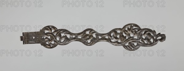 Hinge, unknown author; fittings, openwork strap hinge from a pair: at one end a flat bar bent at a right angle and rolled into a sleeve, the remaining openwork part with a repoussé twig motif, chiseled and engraved; 2nd half of the 17th century; Central Europe; locksmithing., A long, narrow carved object with an openwork, interlacing design running along its length, showing a group of flowing, curved shapes that form circular and looping patterns; the surface appears textured and slightly irregular with a metallic appearance. The colors visible are dark gray, black, and hints of brownish tones, set against a pale gray background.