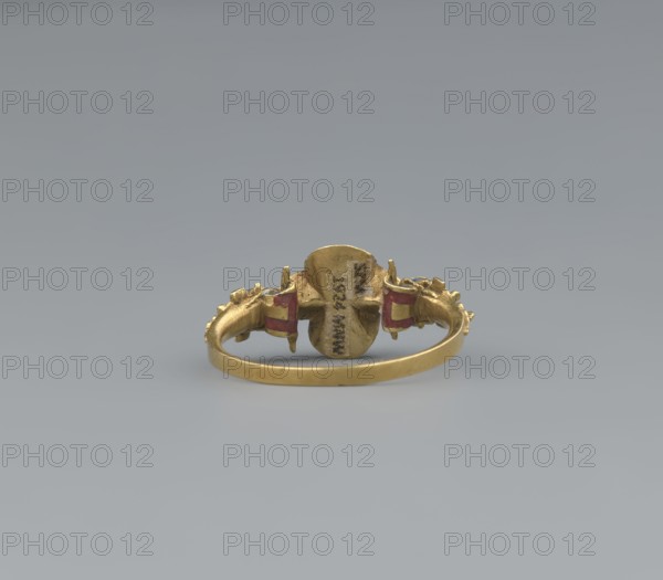 Women's ring, 19th century, Poland; jewelry; height 2.4 cm, width 2.1 cm, A small gold ring shown from the front with a rounded band and an ornate central element flanked by decorative details; the central element is irregularly shaped and slightly convex, while the sides feature sculpted motifs that include small raised forms and curved shapes. The ring shows areas of red pigment on the side decorations and the metal surface has a warm yellow-gold color with subtle variations in tone and a slightly worn, textured finish. The background is a smooth neutral gray gradient.