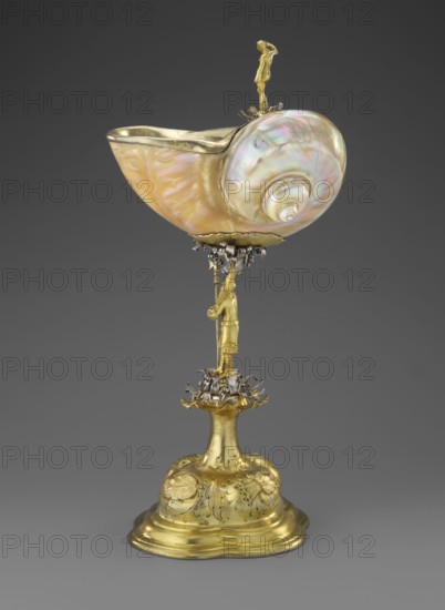 Nautilus Cup. Shell cup on a four-part profiled foot that transitions into a knobbed foot decorated with flowers. The shaft is formed as a figure of a man holding a halberd, standing on a decoration of volute silver ribbon; the motif is repeated at the base of the cup. The rim of the shell is framed in silver and topped with a figure of a nude woman with her right hand raised, probably once holding a now-missing element. Case included. Baroque style, masterpiece; 2nd half of the 17th century, Central Europe. Materials: shell and silver. Subject: men, women, shell shape. Function: vessel. Dimensions: height 37 cm, width 17 cm, depth 18 cm., An ornate object consisting of a large, iridescent shell with pearly shades of pink, green, and cream mounted on a gilded metal stand; the shell bowl shows swirling nacre tones and a smooth glossy surface, with a metallic rim. The stand is gold-colored with sculpted floral and leaf motifs on a rounded base, a fluted stem rising to a decorative platfo...