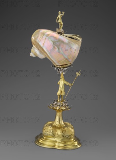 Nautilus Cup, shell cup on a four-part profiled foot that transitions into a knobbed foot decorated with flowers; shaft formed as a figure of a man holding a halberd, standing on a decoration of volute silver ribbon repeated at the base of the cup; rim framed in silver and topped with a figure of a nude woman with her right hand raised, probably holding a missing element; case included. Baroque style, masterpiece. 2nd half of the 17th century, Central Europe. Vessel; height 37 cm, width 17 cm, depth 18 cm., A decorative object composed of a large iridescent shell with pearly pink, green, and gold tones mounted on a metallic support, topped by a small gold figure standing on the shell, above a group of ornate silver and gold embellishments; below the shell a gold humanoid figure holds a long staff with a pointed end and stands on a floral-like gold and silver platform, all rising from a gold base with rounded edges and embossed leaf-like decorations, set against a plain gray background.