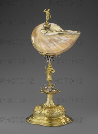 Nautilus Cup. Shell cup on a four-part, profiled foot that transitions into a knobbed foot decorated with flowers. The shaft is a figure of a man holding a halberd, standing on a decoration of volute silver ribbon; the motif is repeated at the base of the cup. Rim framed in silver and topped with a figure of a nude woman with her right hand raised, probably holding a missing element. Case included. Baroque style, masterpiece. 2nd half of the 17th century, Central Europe. Vessel. Height 37 cm; width 17 cm; depth 18 cm. Men; women; shell shape., A decorative object consisting of a large iridescent shell with pearly hues of pink, cream, and green mounted on an ornate metal stem; the shell is partially lined with a shiny golden rim and topped by a small golden human-like figure standing on a floral mount. The stem features a golden human-like figure holding a long staff and stands above a complex floral and scrollwork cluster in mixed metallic tones of gold and silver. The base is a rounde...