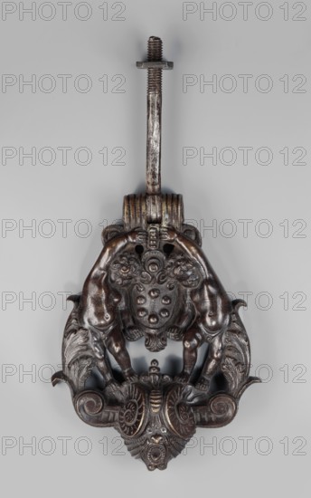 Renaissance door knocker with the Medici coat of arms, Renaissance style. Bronze door knocker with a base in the form of a mask with horns in the form of volutes, on which stand two naked putti supporting the Medici coat of arms. The figures are framed on the outside by acanthus tendrils growing from the volutes. A movable pin for mounting the door knocker is placed above the coat of arms. A similar object is located in the Museum of Decorative Arts in Prague. Ryszard Bobrow noted the popularity of this model in the 19th century, especially in Paris, where cast-iron knockers of similar shapes can still be found on the doors of tenement houses. 16th century, Italy, Venice; door fittings; height 21 cm, width 18 cm., A dark metallic decorative object with a long vertical rod topped by a threaded bolt and flat nut, connected to an ornate sculpted plate featuring a pair of stylized animal figures facing inward, their muscular bodies and limbs forming a symmetrical composition around a centr...