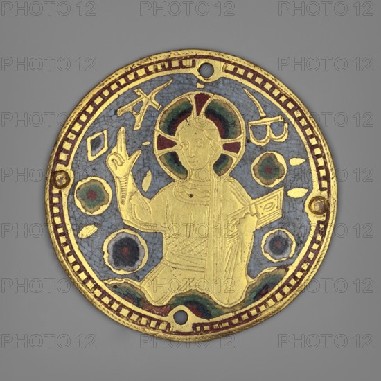 Fragment of the cover of a liturgical book?, enamel work, 19th century, France, Limoges; plaque, diameter 7 cm, A round decorative medallion with a central figure in gold against a blue background, the figure seated and holding an open book in one hand while the other hand is raised; the figure has a halo with green and red segments and wears a robe with engraved lines, and there are several circular floral-like motifs in green and red around the figure; the medallion edge is gold with a band of red rectangles and has a few small holes, set against a gray backdrop.