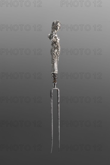 Fork. Two-pronged steel fork with a silver handle in the form of a full-plastic female herm. Unmarked. 2nd half of the 17th century, France . Cutlery. Height 19.5 cm; weight 5 g., A metallic two-pronged fork with a decorative handle shaped like a female bust and ornate scrolling patterns, shown against a gradient gray background; the object displays shades of silver and gray with areas of darker patina and slight brownish discoloration on the metal, and the background shifts from lighter gray at the top to darker gray at the bottom.