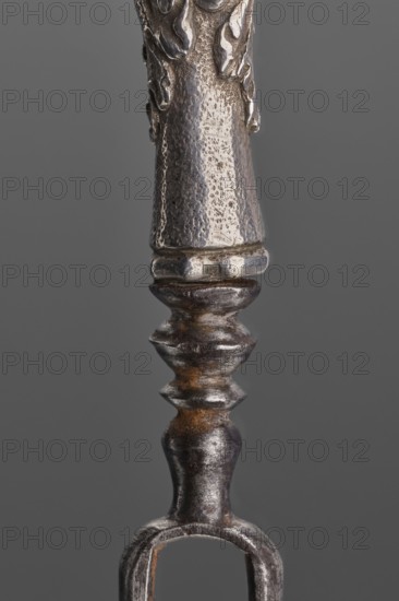 Fork. Two-pronged steel fork with a silver handle shaped as a full-plastic female herm. Unmarked. 2nd half of the 17th century, France . Height 19.5 cm; weight 5 g., Repossession; cutlery; function: dining utensil., A close-up vertical view of a metallic object with a textured surface showing a few rounded and ridged segments; the metal appears mostly silver-gray with areas of darker gray and brownish rust tones; the background is a uniform mid-gray.