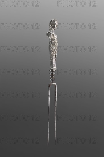 Fork. Two-pronged steel fork with a silver handle shaped as a full-plastic female herm, decorated with acanthus in the lower part. Unmarked, 2nd half of the 17th century, probably France. Cutlery; height 19.5 cm, weight 5 g., A metallic two-pronged fork with a decorative handle shaped like a stylized human figure, the handle showing ornate carved details and flowing motifs; the metal appears predominantly silver with areas of darker tarnish and slight rust on the prongs, set against a gradient gray background.