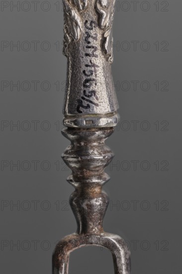 Fork. Two-pronged steel fork with a silver handle shaped as a full-plastic female herm, decorated with acanthus in the lower part. Unmarked, 2nd half of the 17th century, France, cutlery, height 19.5 cm, weight 5 g., A close-up of a metallic object with a textured surface showing a tapered top section and a turned, bulbous central shaft leading to a forked lower end, displaying a mix of silver, gray, dark brown, and rusty orange tones against a neutral gray background.