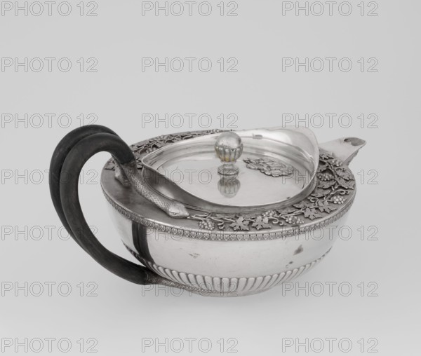Teapot, Rundell Bridge & Rundell fl. ca. 1788–1845 goldsmith's firm; Scott, Digby & Smith, Benjamin fl. 1802–1807 goldsmiths/sellers. Tea vessel; hemispherical body on a low cylindrical pedestal, lower body decorated with radial curls, upper edge with a band of acanthus leaves. Hinged lid with raised collar bearing an applied vine branch with clusters. Double handle of blackened wood set in caps upper caps shaped as snake heads, lower as tails. Applied Czartoryski coat of arms on a paludament with labry and the motto Badz co sie jest; also bears Pogon Ruska coat of arms, rank crowns, princely mitre, mottos heraldry, paludamentum, labry. Ornament: acanthus leaves, vines, snake iconography, clasp. Inscription/motto: Badz co sie jest. Date: 1803/1804, London, England. Materials: silver with applied ornament, blackened wood handle. Dimensions: height 13 cm; width 24.6 cm; depth 17.2 cm; weight 1002 g., A reflective metal bowl-shaped vessel with an ornate lid sits against a plain light gray...