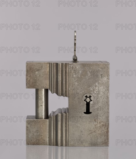 Padlock, 19th century. A cuboid-shaped padlock with the upper part topped by a cornice; on each side two cuboids are connected by a cylindrical rod acting as an earring. After unlocking, the side wall together with the earring is pulled out, aided by an openwork hinged handle. The keyhole is horizontal on the longer wall. Materials/function: locksmithing; height 12.6 cm, width 12.7 cm, depth 6.2 cm., A metallic rectangular object with a weathered, grayish-silver surface sits centered against a pale lavender-gray background; the object has a hollow vertical channel on the left with a smooth cylindrical rod spanning between the top and bottom sections, and the central edges of the channel show a series of scalloped ridges; the right face of the object is flat and worn, with a dark silhouette shaped like a keyhole that includes a star-like upper opening and a narrow stem; a thin, slightly curved metal loop rises from the top center, attached by a small decorative rivet, and the object cas...